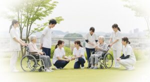 特養の庭で、高齢者、介護士、ボランティアが笑顔で交流している様子。多世代交流と地域連携が感じられる明るい屋外の風景。